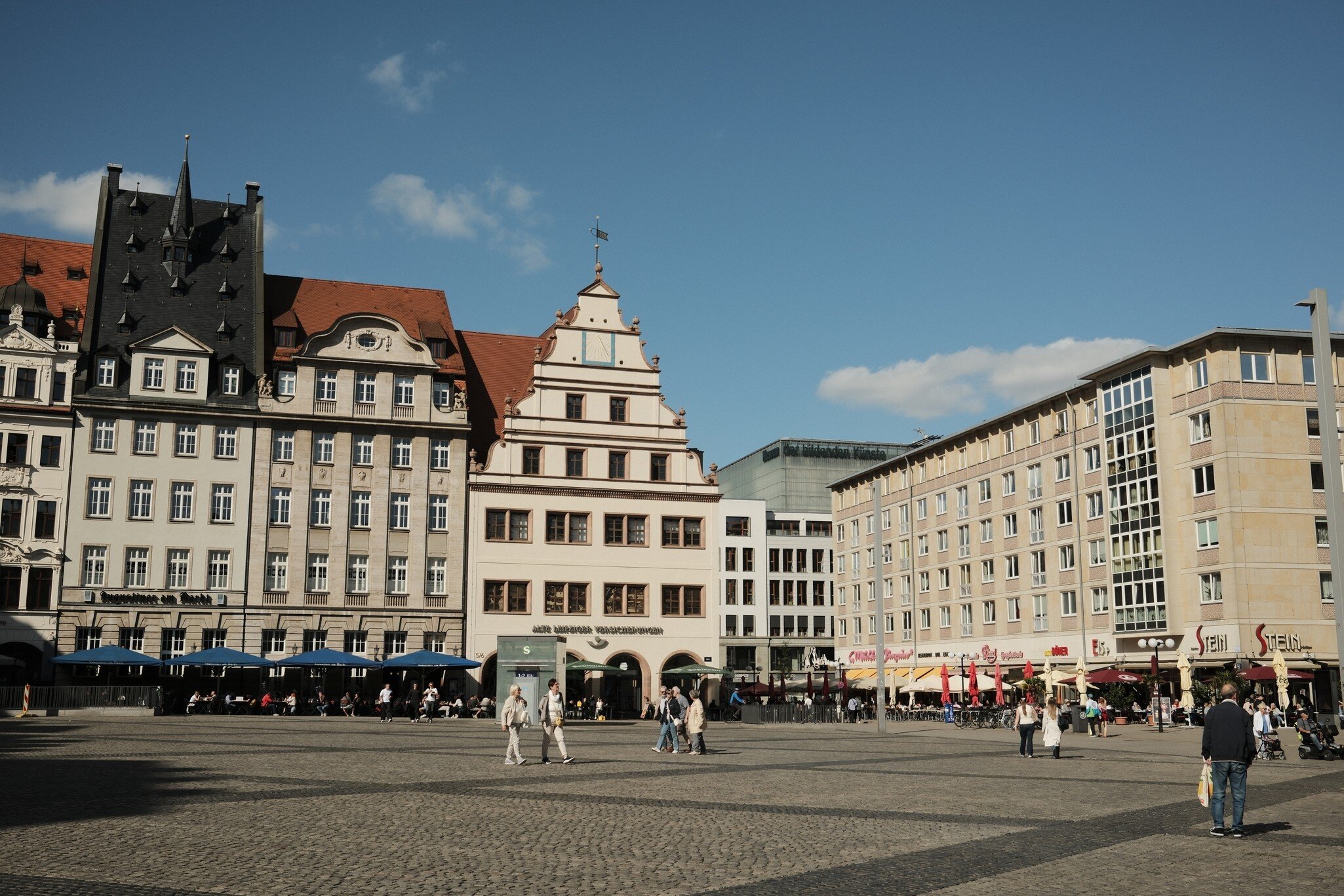 People cross the cobbled Markt square in Leipzig, Germany, lined with gabled historic buildings and cafe umbrellas