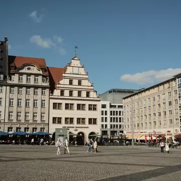People cross the cobbled Markt square in Leipzig, Germany, lined with gabled historic buildings and cafe umbrellas