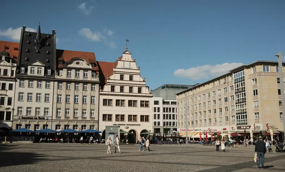 People cross the cobbled Markt square in Leipzig, Germany, lined with gabled historic buildings and cafe umbrellas