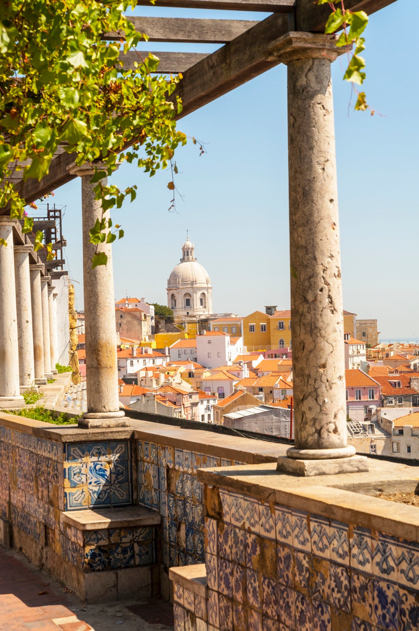 Stone columns and a vine-draped pergola above blue-and-white tiled walls, with orange rooftops and a white dome beyond.
