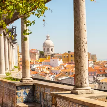 Stone columns and a vine-draped pergola above blue-and-white tiled walls, with orange rooftops and a white dome beyond.