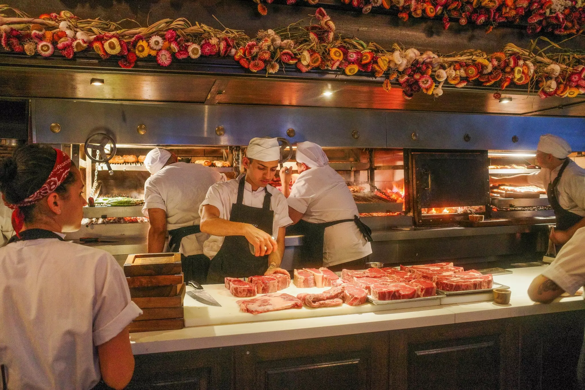 A chef seasons raw cuts of red meat on a counter at a restaurant. Other chefs and workers staff the kitchen around him.
