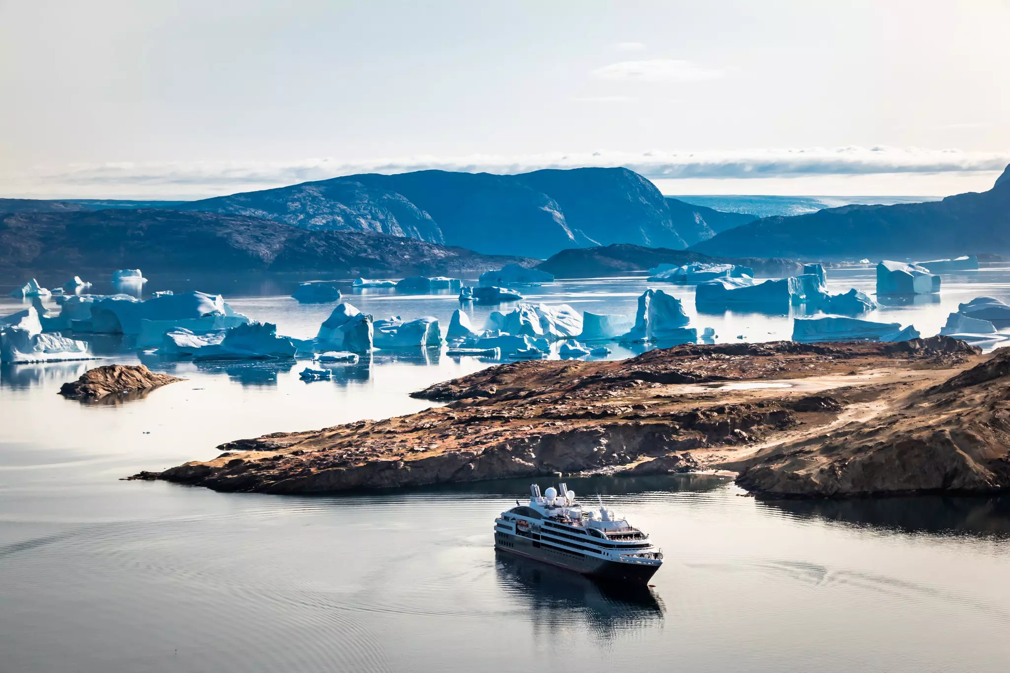 A cruise ship sails beyond a cove with icebergs in bluish light in Greenland.