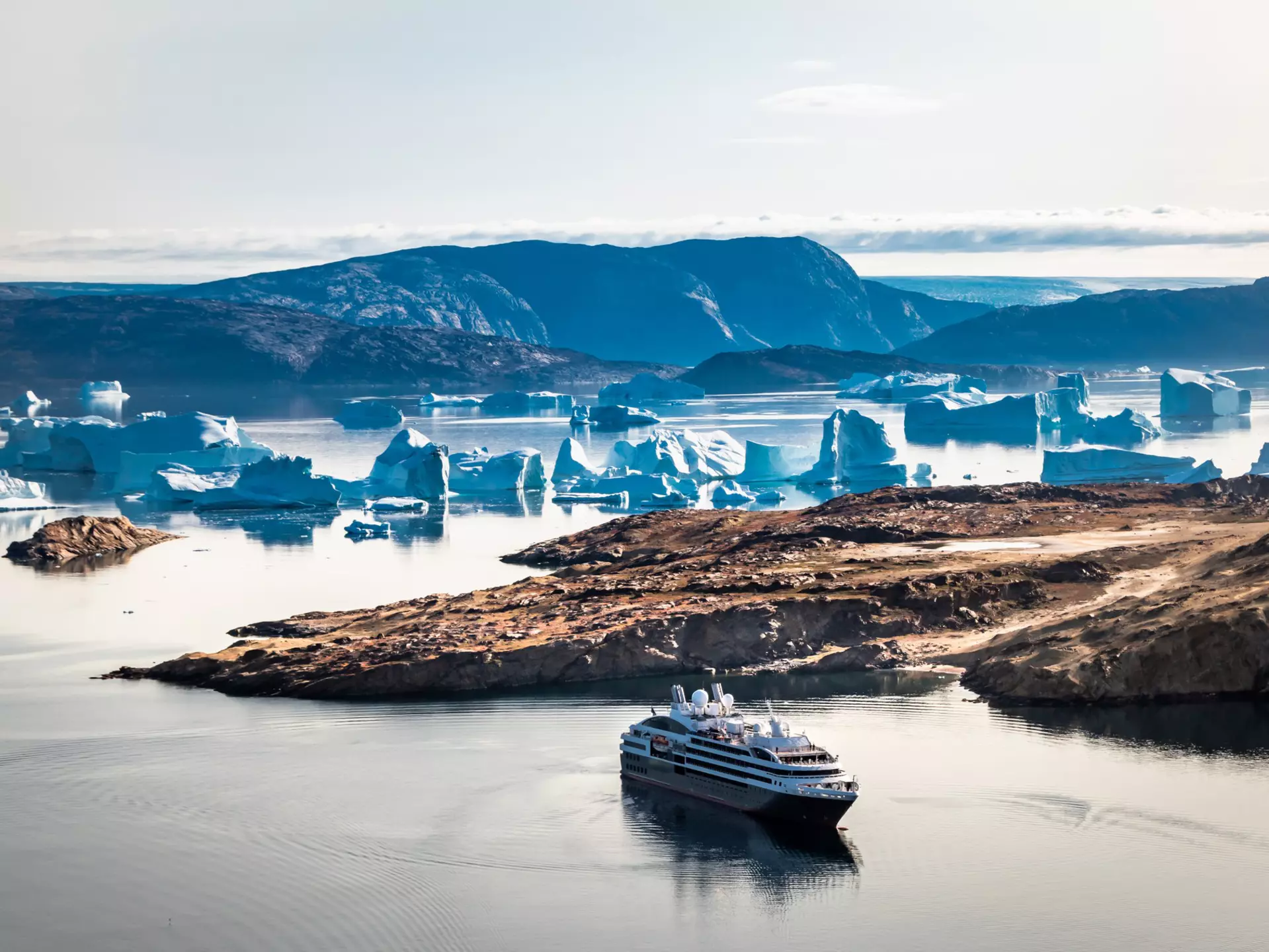 A cruise ship sails beyond a cove with icebergs in bluish light in Greenland.