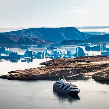 A cruise ship sails beyond a cove with icebergs in bluish light in Greenland.