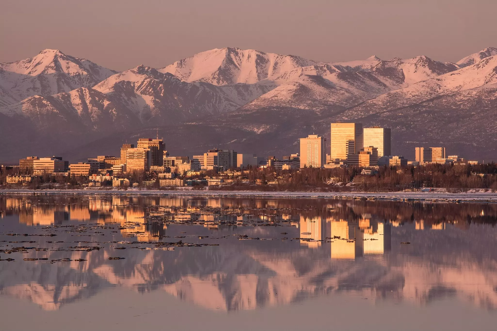 The skyline of a city is framed between a bay that reflects its buildings and snow-capped mountains in the distance.