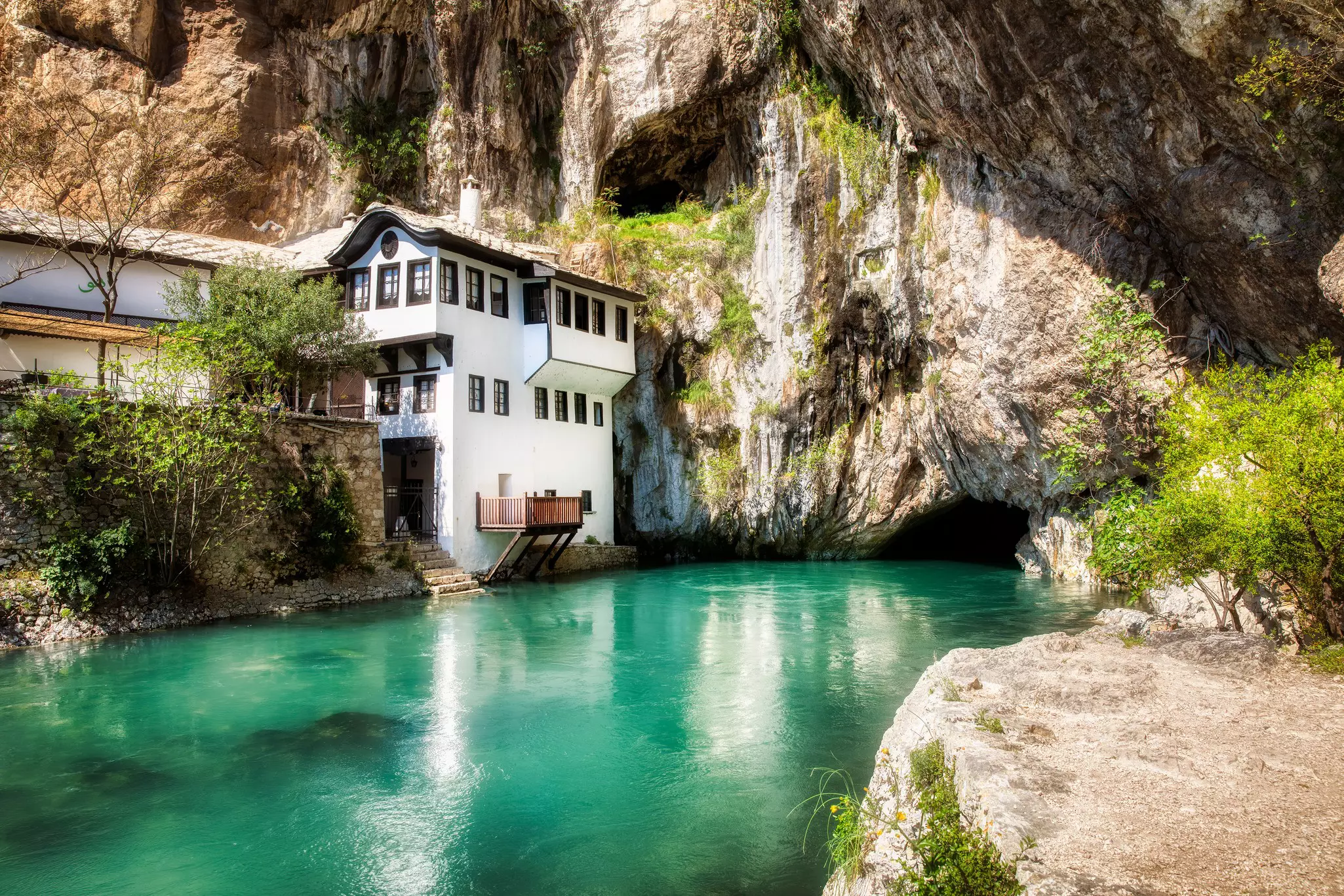 A small white building built into the foot of a cliff right near a turquoise river.
