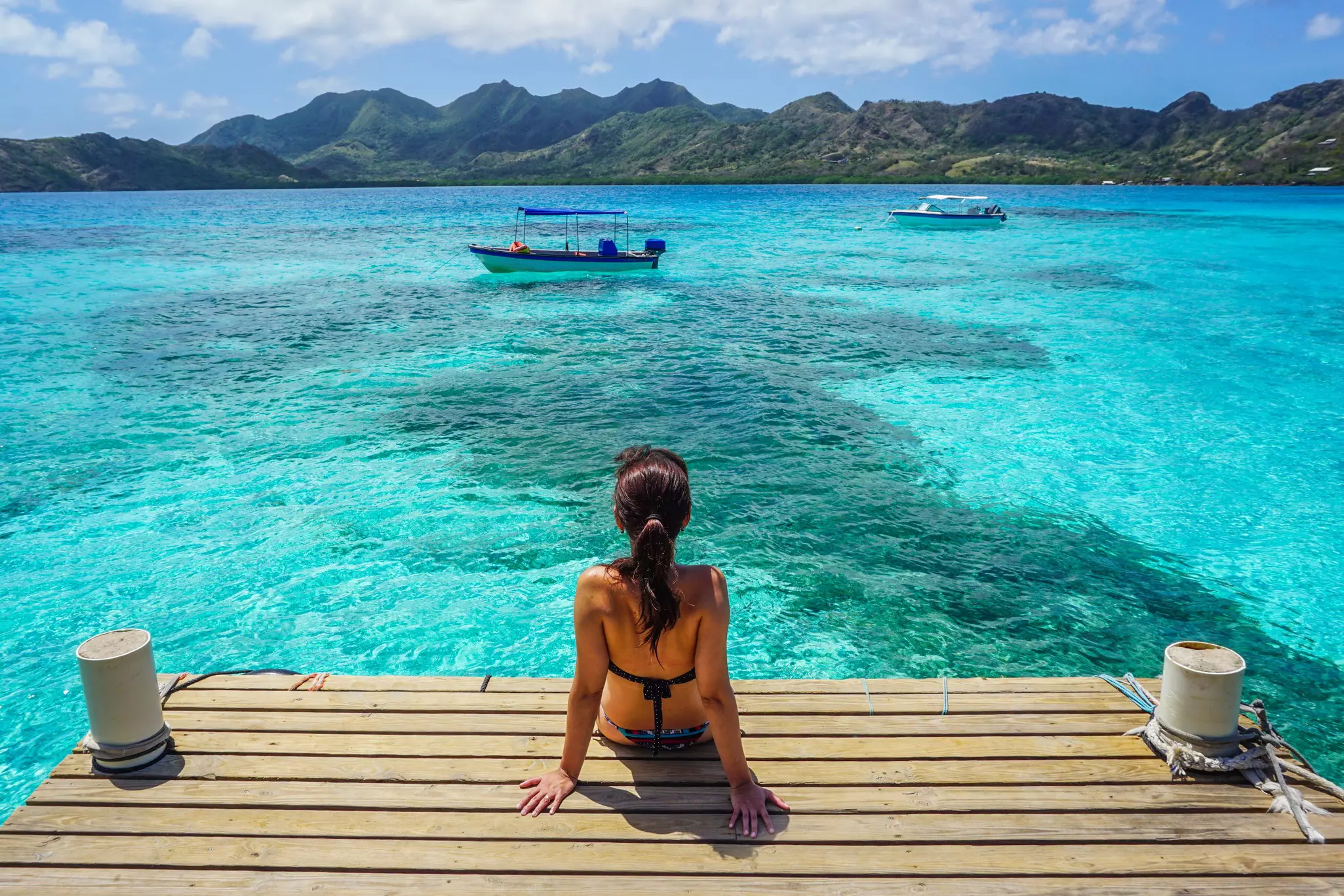 A woman sitting on a dock is seen from behind, as she looks out at a boat in the blue waters off a tropical island.