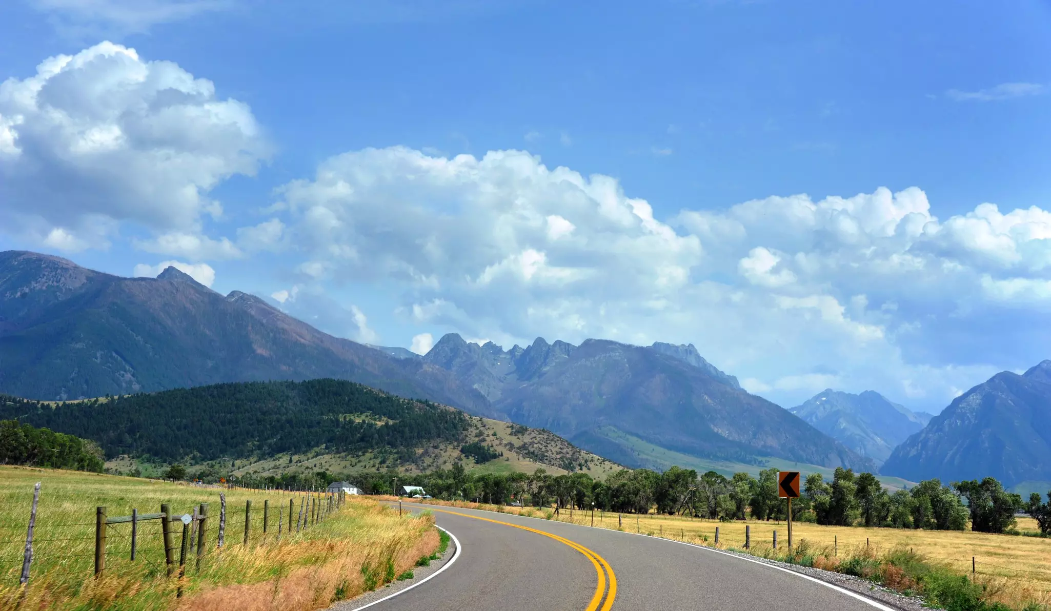 A single-lane highway curves through grassland heading towards mountain peaks.