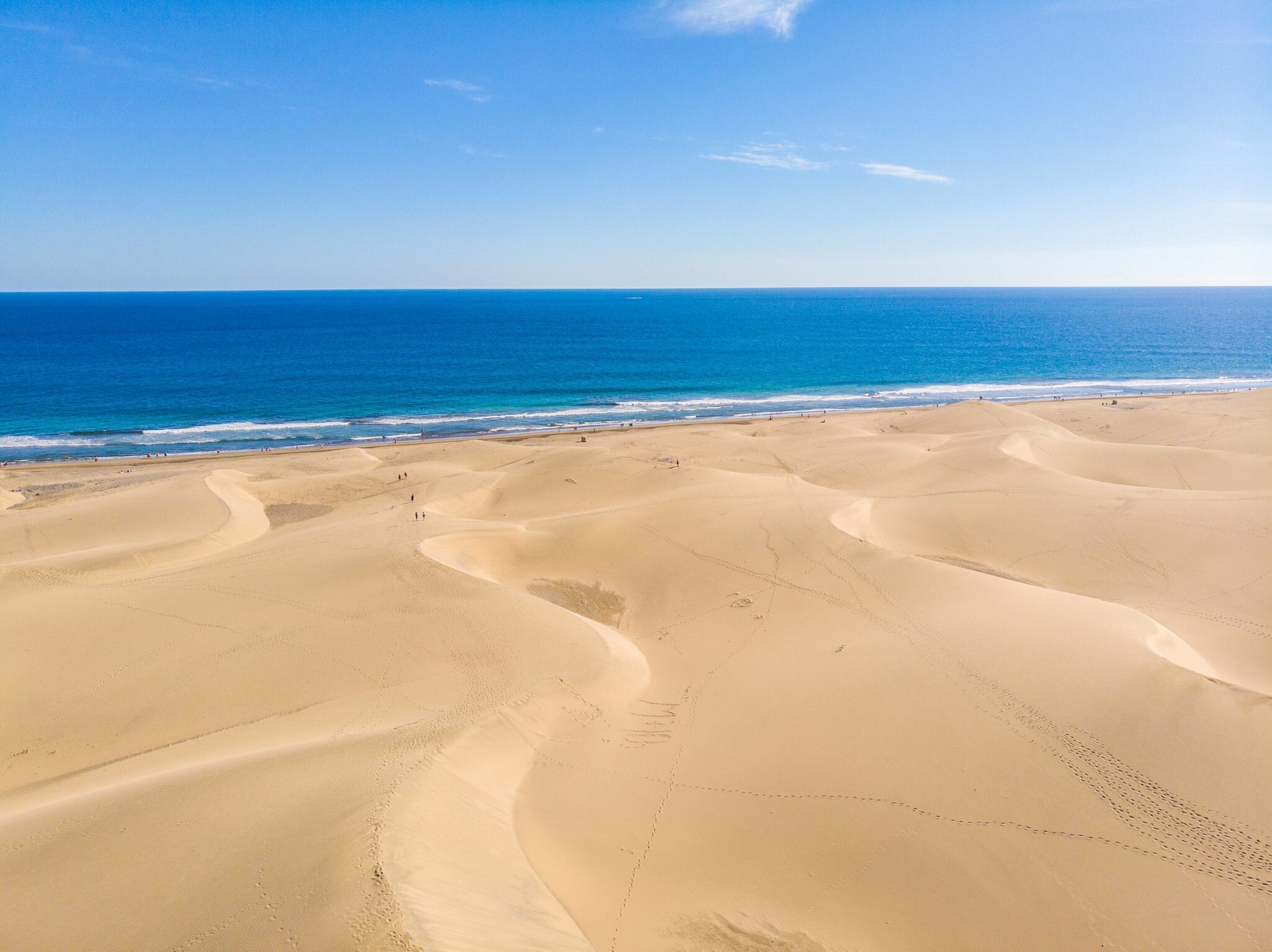 Rippled golden dunes with footprints stretching to a strip of sand, white surf and deep blue ocean under a clear sky