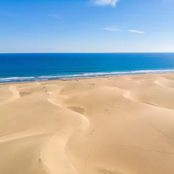 Rippled golden dunes with footprints stretching to a strip of sand, white surf and deep blue ocean under a clear sky