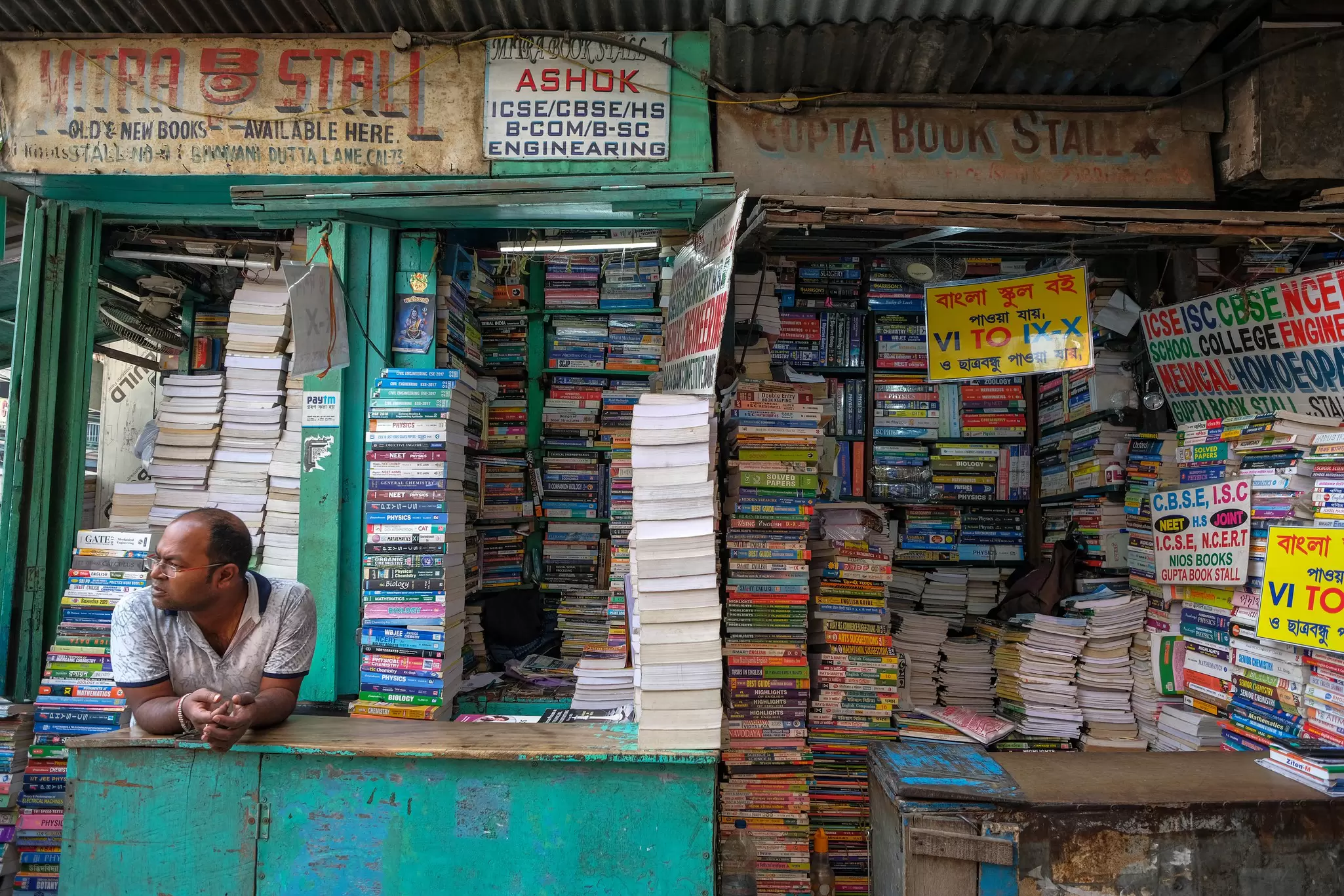 A man leans over a counter; a stall filled with books is behind him.