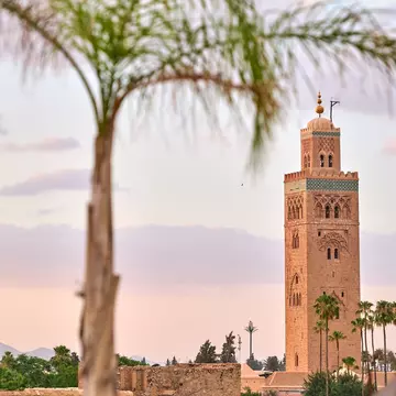 Sandstone minaret with turquoise tilework and a gold finial, framed by blurred palm fronds against a pink sky.
