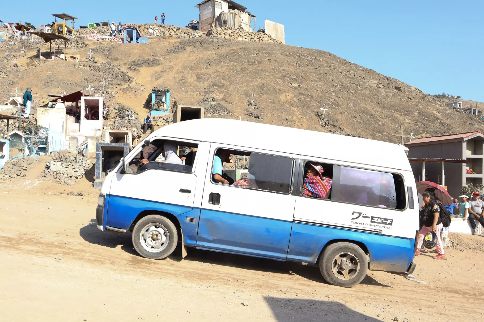 A white van packed with passengers drives up a sloped road in a cemetery.