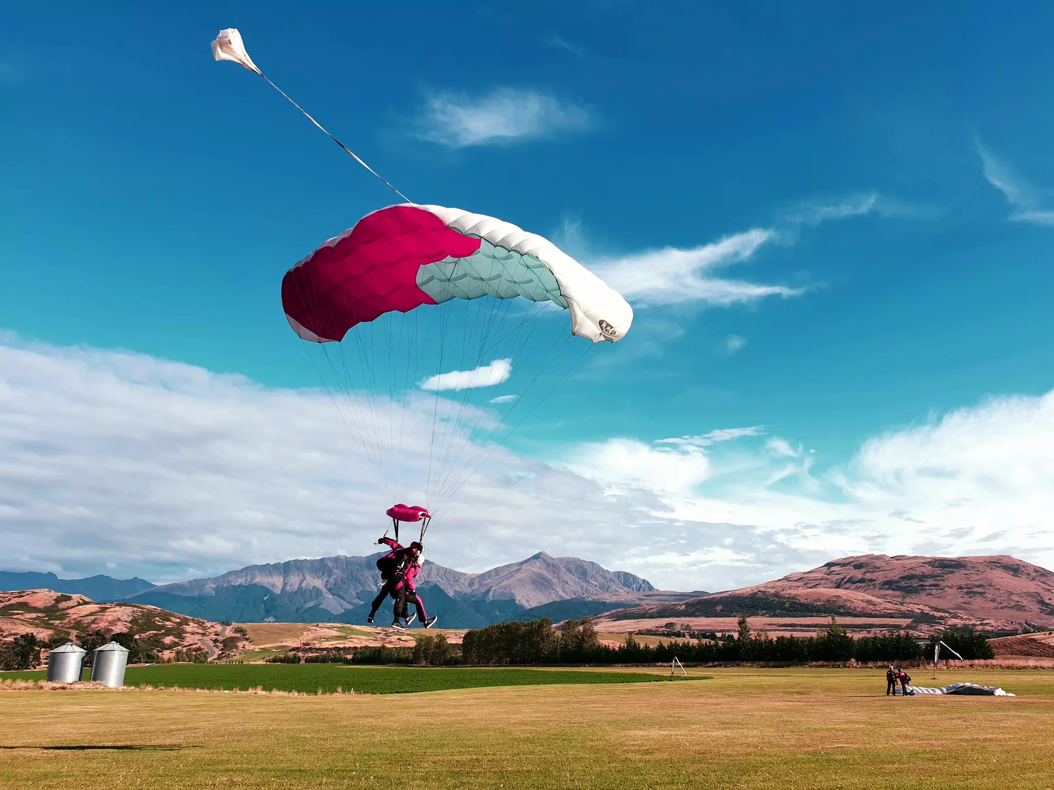 A skydiver, strapped to an instructor, come into land at an airfield with their red and white parachute fluttering above them