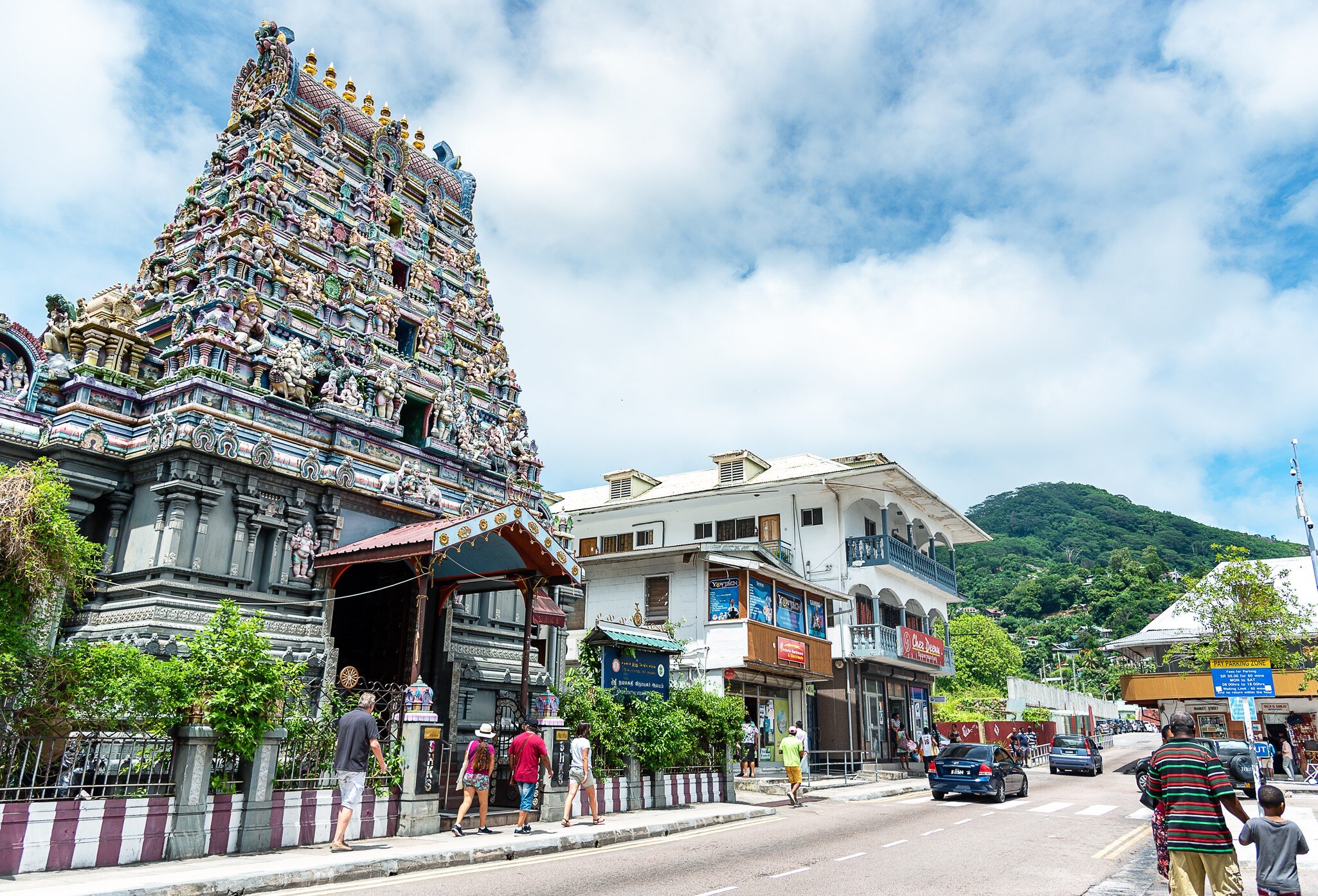 Tall tower covered in carved deity figures beside a white two-storey building, people walking past and green hills behind