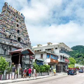 Tall tower covered in carved deity figures beside a white two-storey building, people walking past and green hills behind