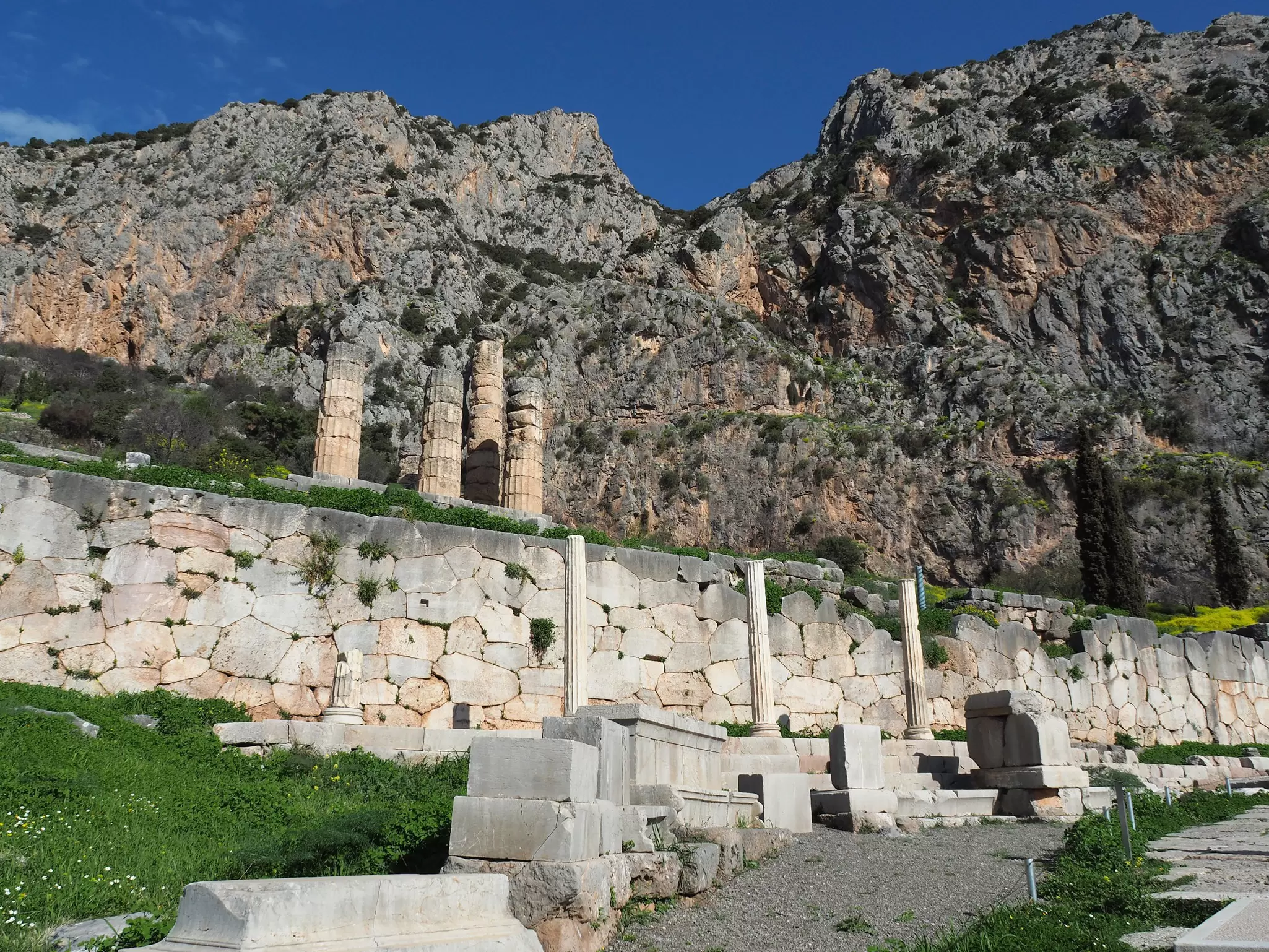 A view Along The Sacred Way in Delphi, Greece. The white walkway is lined by stone ruins of temples and other structures.