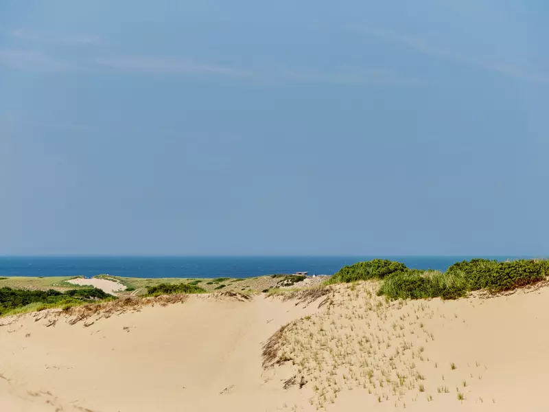 Dunes of beige sand with tufts of grass in front of a low horizon where blue sea and sky meet.