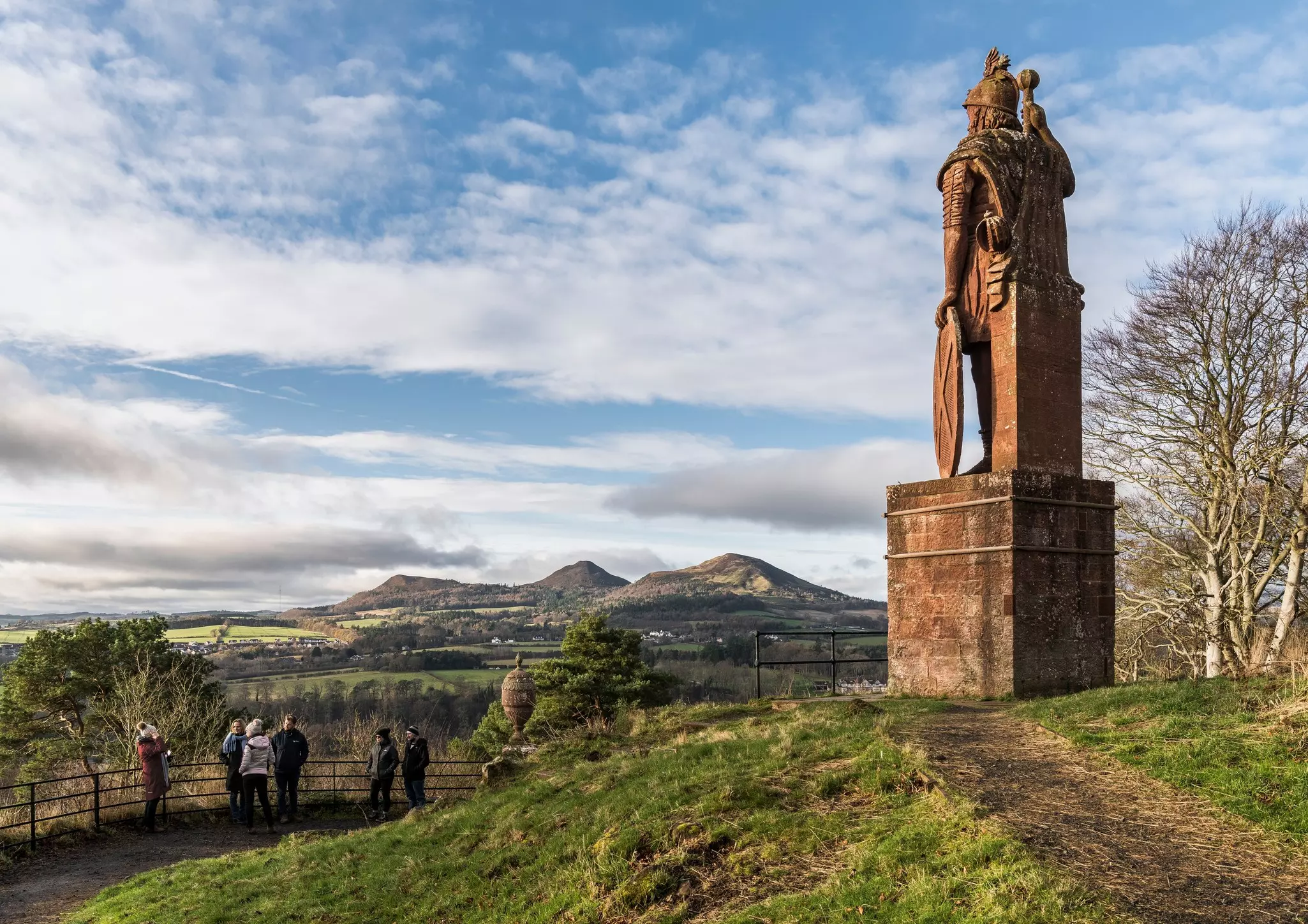 People pause at a viewpoint in a hilly rural area near a tall statue of a man looking into the distance