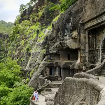 Two people in white walk a path below carved cave entrances cut into a cliff at Ajanta, India, surrounded by green trees