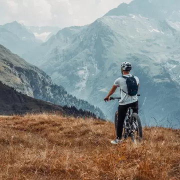 A cyclist with a helmet stands on dry grasses at a mountain overlook.
