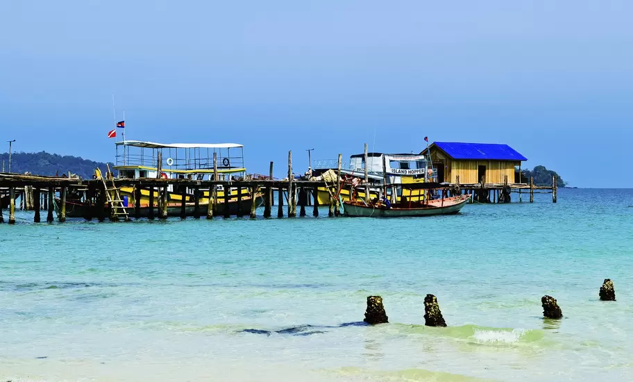 Wooden pier on Koh Rong, Cambodia, with tour boats, an Island Hopper ferry and a blue-roofed hut over clear turquoise water