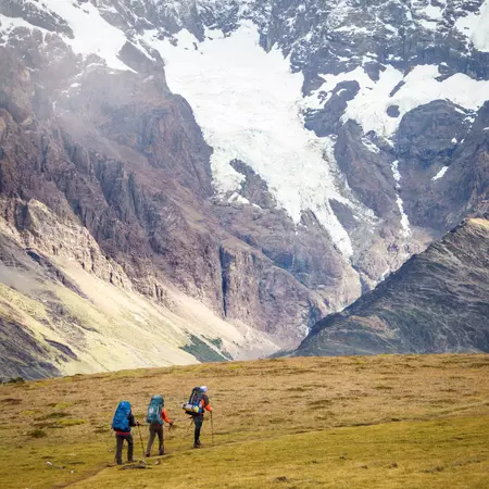 Three hikers walk over a grassy path; patches of snow on a rocky mountain are in the background.