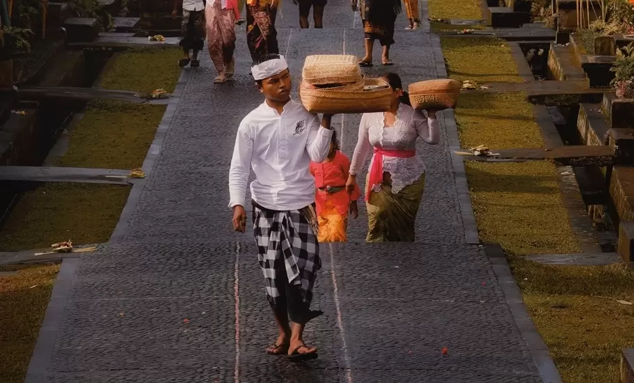 Balinese villagers carrying offering baskets on their heads along a stone lane lined with arching penjor poles, Indonesia