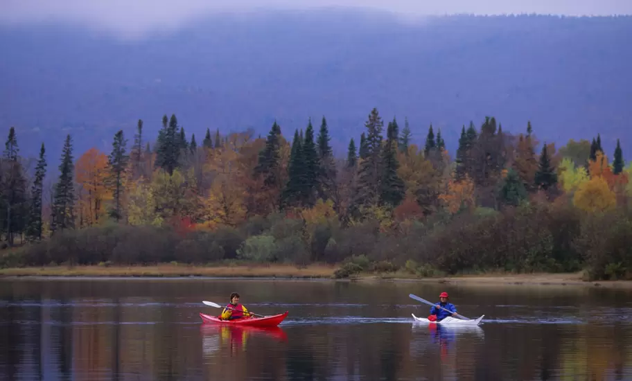 Two kayakers paddling a calm lake before a shoreline of autumn foliage and mist-shrouded forested hills