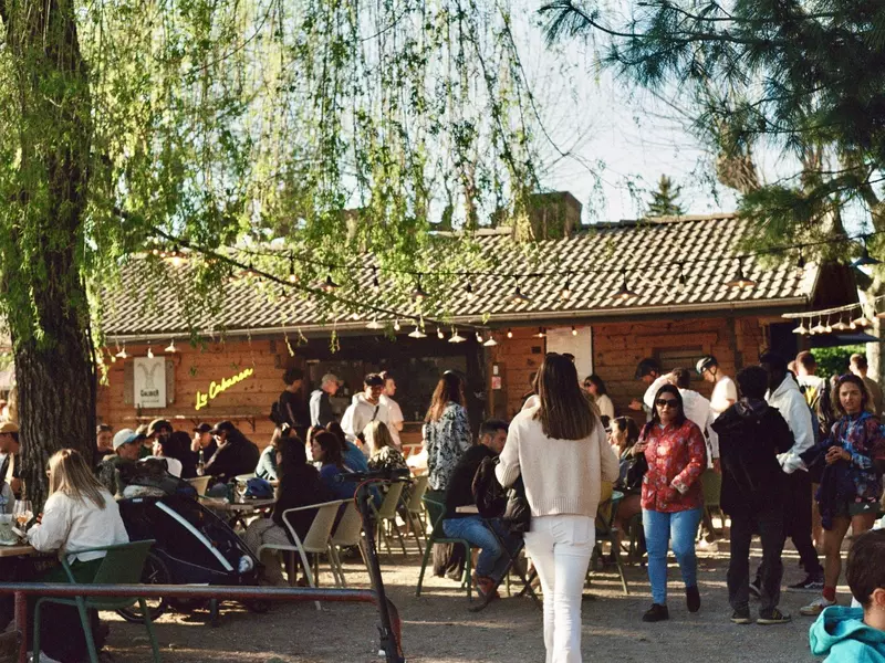 People at and around picnic tables outside at a bar in France; there is a large tree providing shade, and a wood building is in the background.