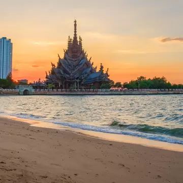 Ornate wooden temple with tall spires on a seawall beside a sandy beach, high-rise towers and an orange sky behind