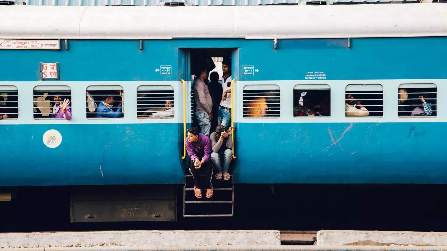 A blue train carriage with slatted windows and many passengers sat at seats inside. Several people crowd round the open door with two sat on the steps.
