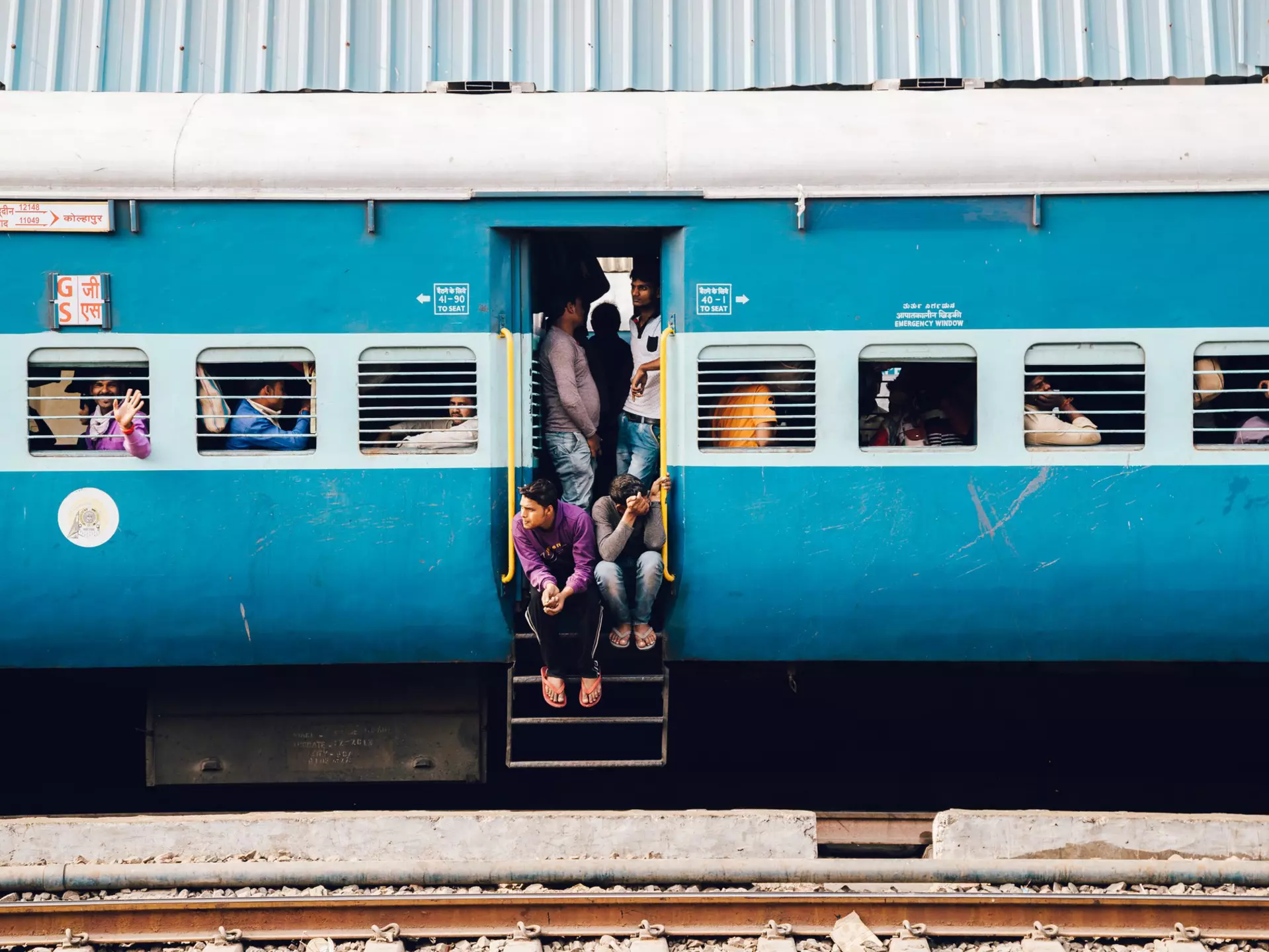 A blue train carriage with slatted windows and many passengers sat at seats inside. Several people crowd round the open door with two sat on the steps.