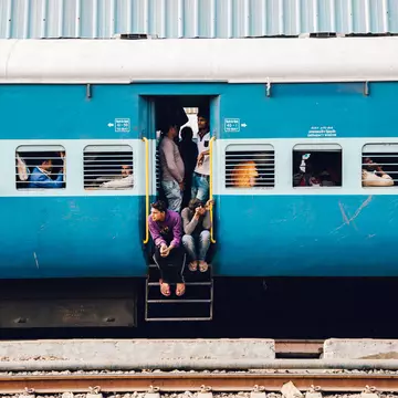 A blue train carriage with slatted windows and many passengers sat at seats inside. Several people crowd round the open door with two sat on the steps.