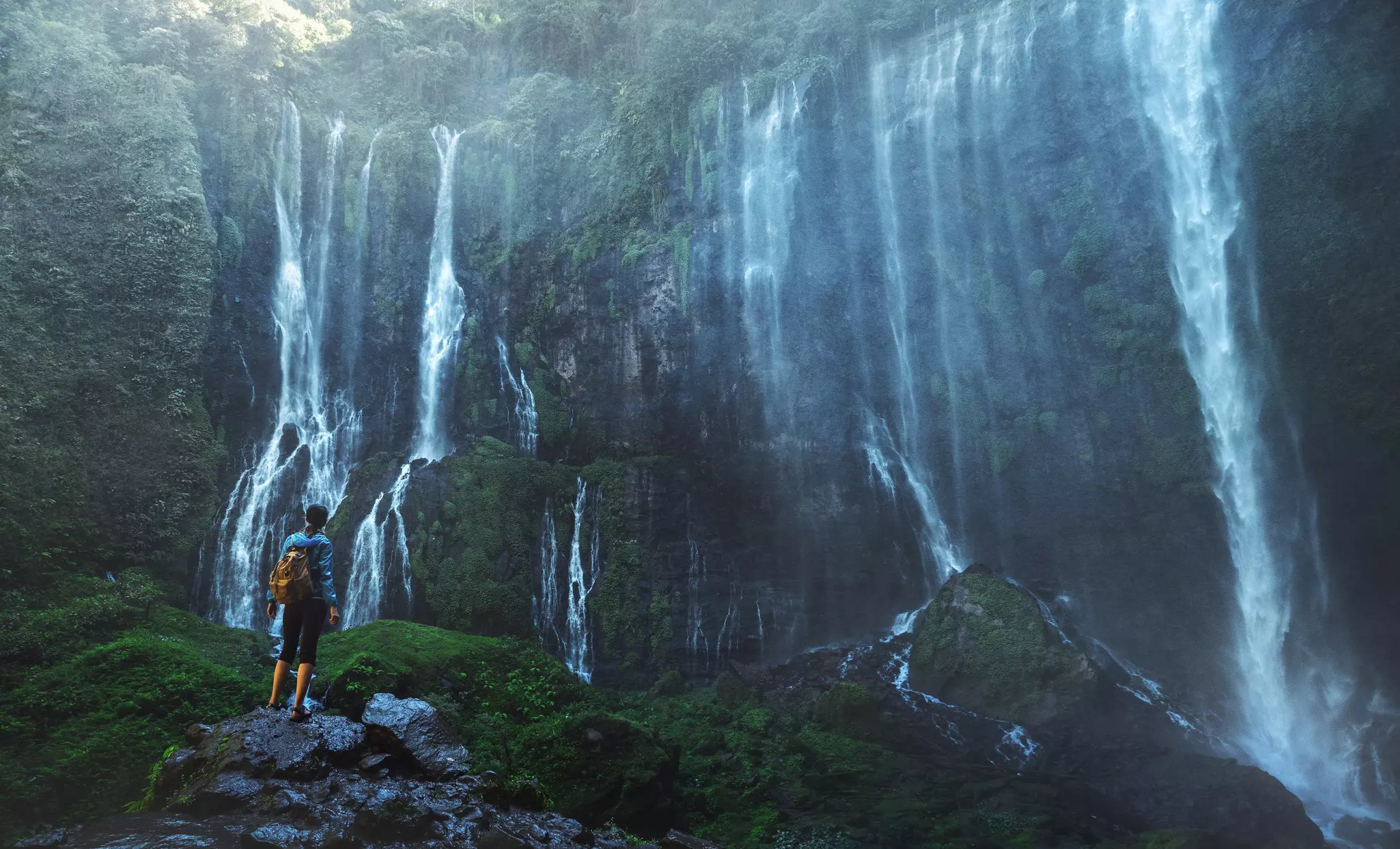 A man stands on a rock and gazes up at a huge waterfall with hundreds of streams
