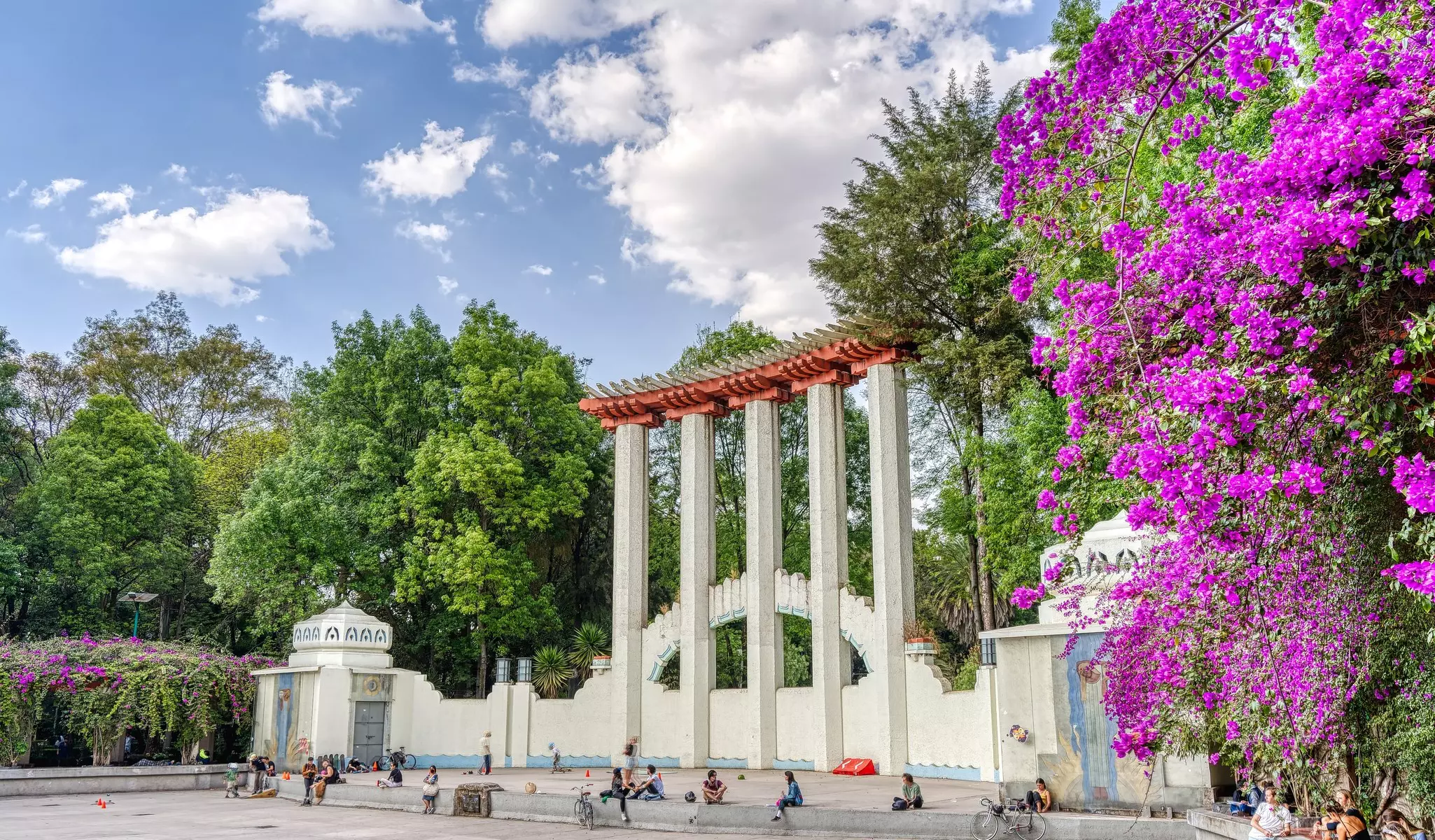 People sitting in a park in Mexico City in front of a giant row of columns and cascading flowers.