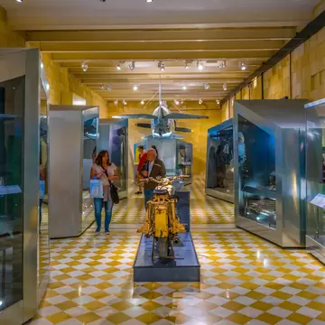 A vaulted gallery with a chequered tiled floor, glass cases of uniforms and helmets along both walls, a military motorcycle on a plinth and a fighter aircraft suspended at the far end.