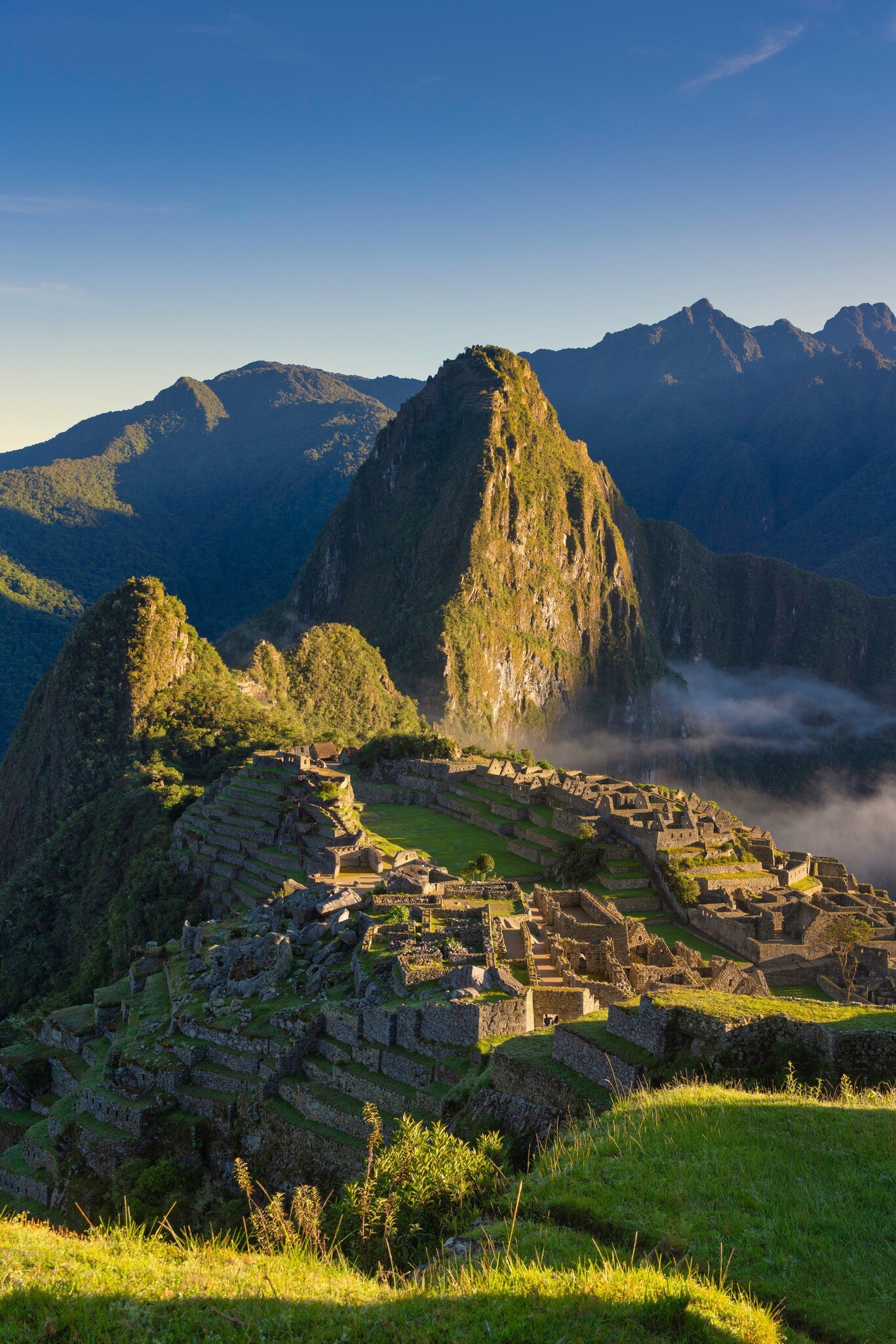 Stone ruins and terraces of Machu Picchu below the steep green peak of Huayna Picchu, with low cloud in the valley.