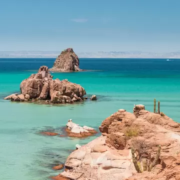 Lone stand-up paddleboarder crossing turquoise shallows past cactus-topped red rocks, Isla Espiritu Santo, Baja