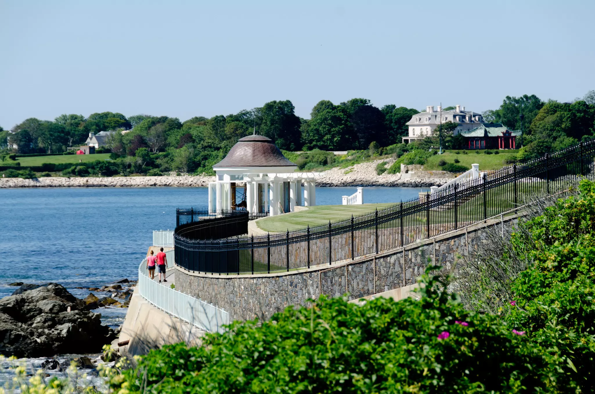 Two people walking on a path by the water; there is a gazebo ahead and a large house with a green lawn.