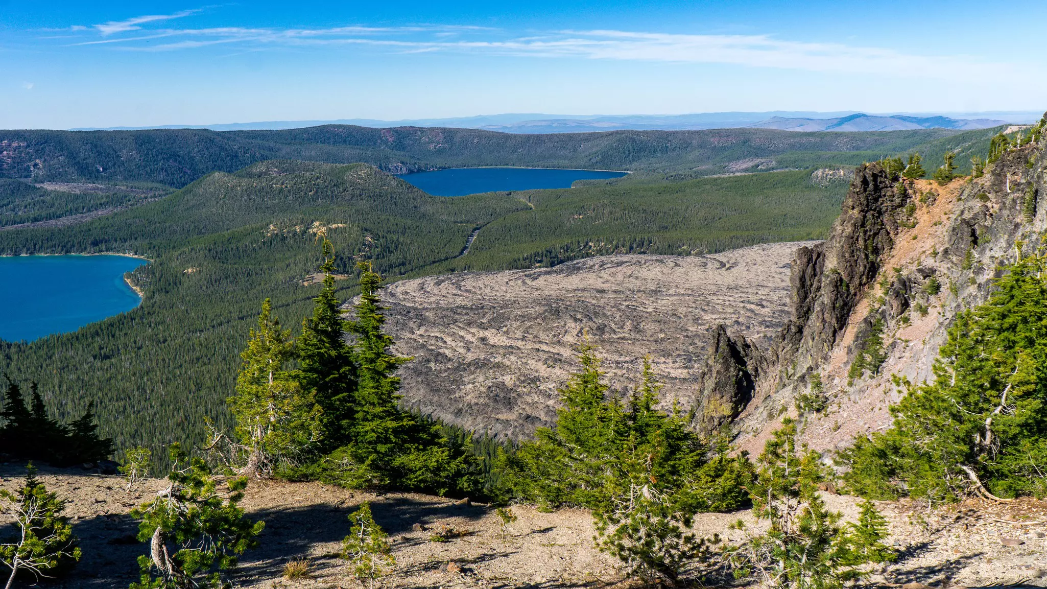 Aerial view of two caldera lakes surrounded by forest and an obsidian field on a sunny day.