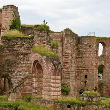Arched brick and sandstone walls of the ruined Roman imperial baths in Trier, topped with grass under a cloudy sky