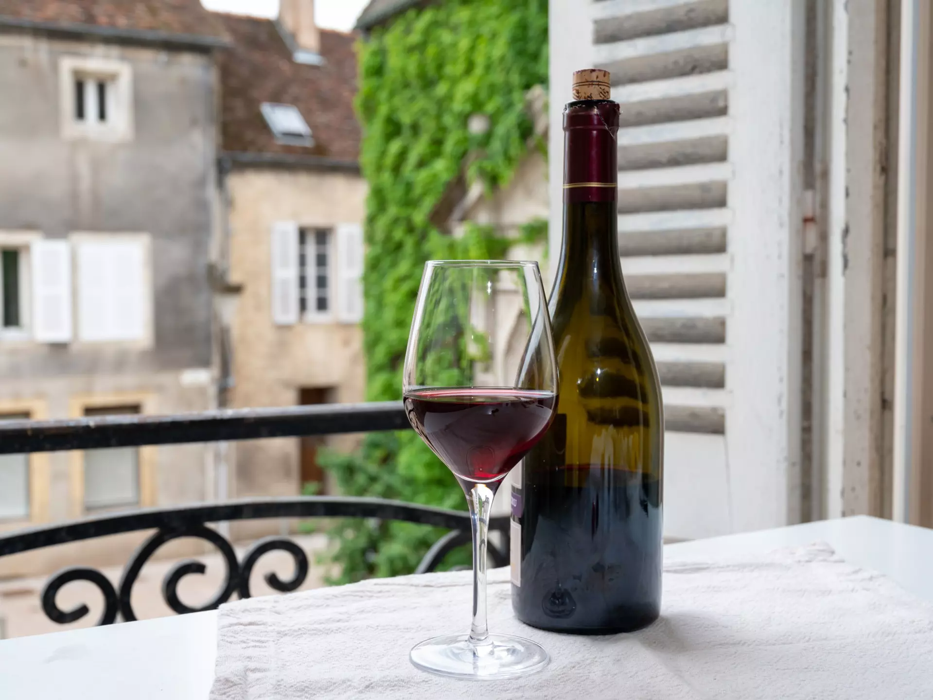 A bottle of red wine and wine glass on a table near a balcony of a historic French building