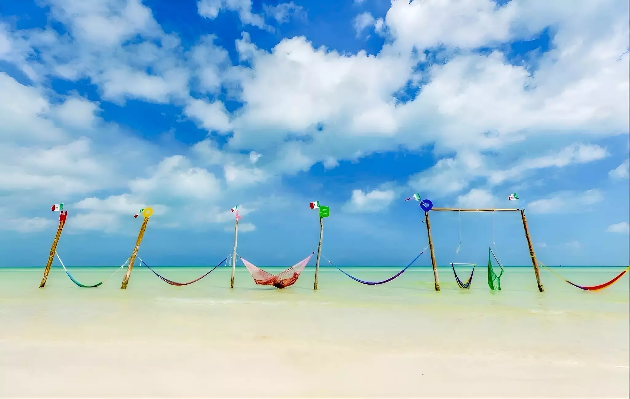 A line of seven multicolored hammocks attached to posts set in shallow turquoise water.