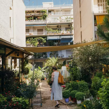 A woman walks through a narrow alleyway with plants for sale on either side and a residential building in the distance.