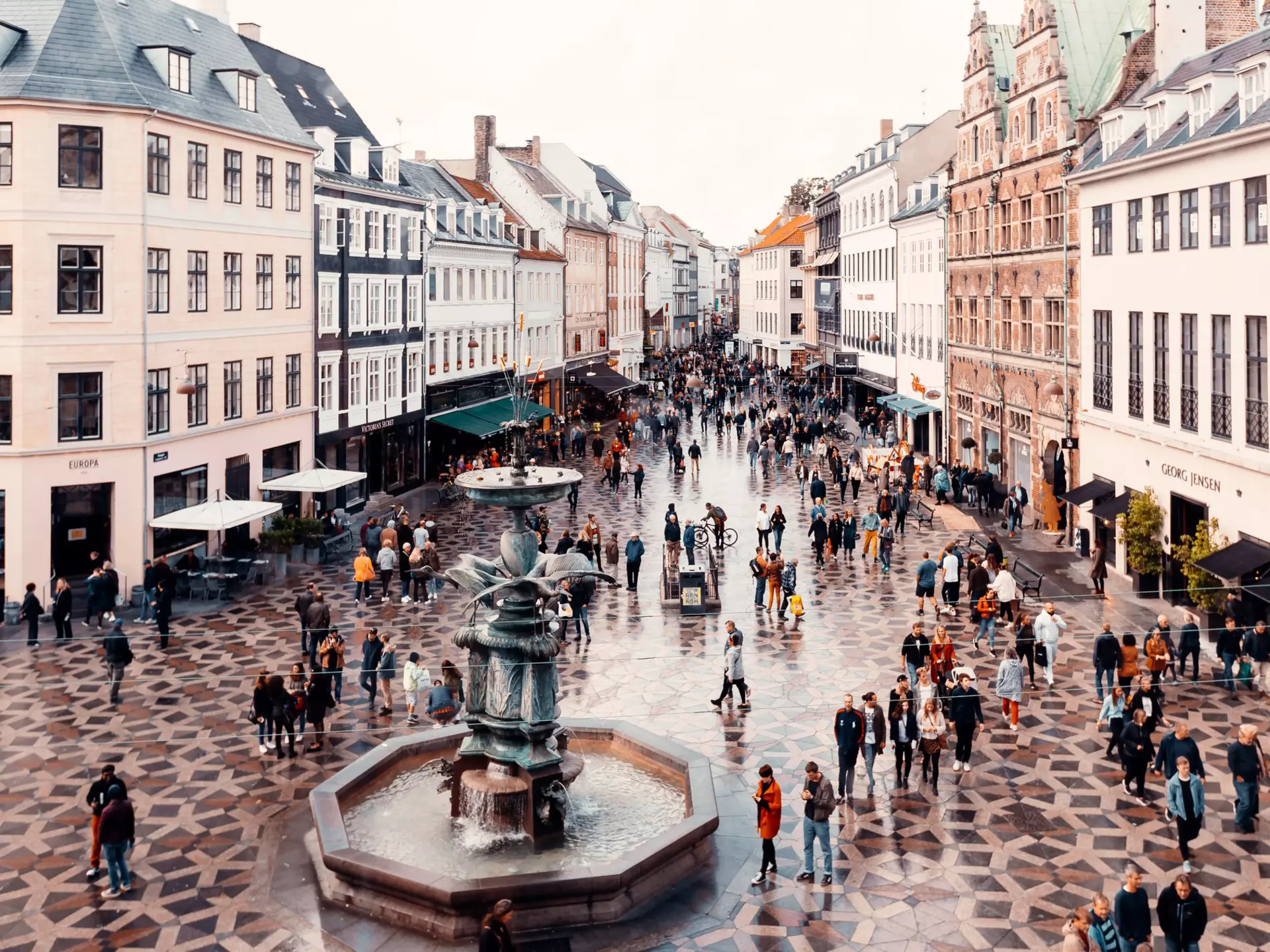 A pedestrianized shopping area with many people walking along the patterned street lined with stores.