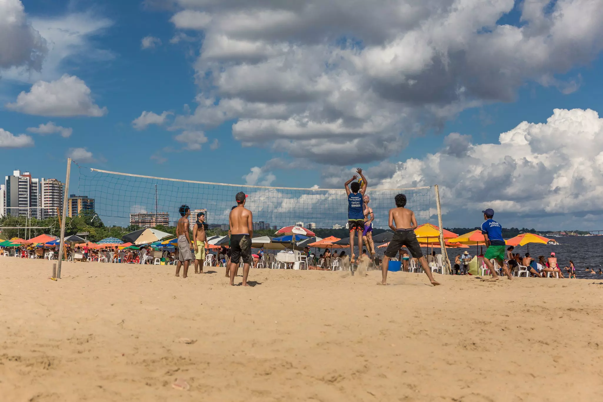 Group of local Brazilians play beach volleyball on the urban Ponta Negra beach in the Amazon while people relax sitting on chairs under sun umbrellas in the background