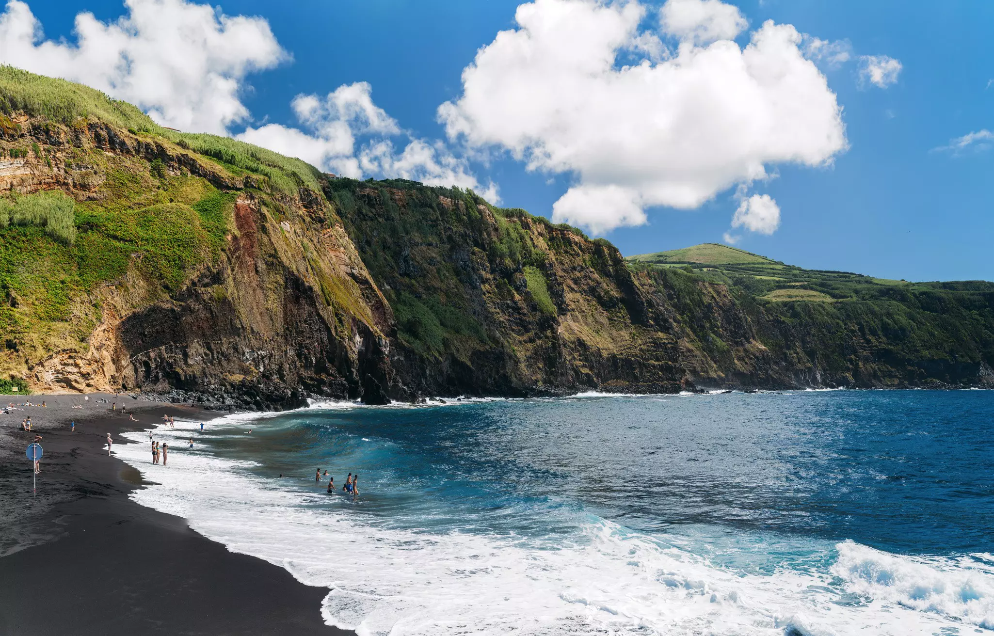 Waves lap a black-sand beach, with people swimming in the surf. Tall cliffs with green vegetation plunge toward the water leading up to the beach.l