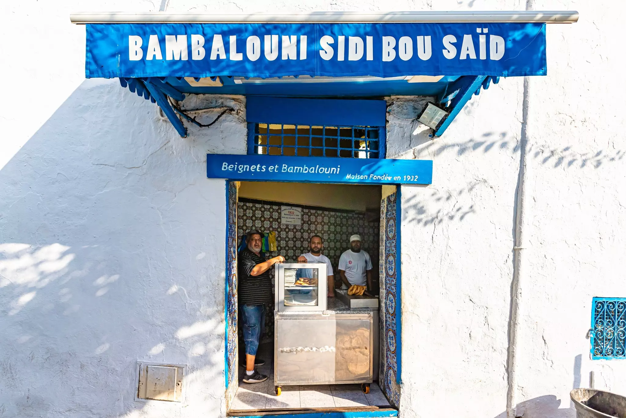 Three men stand inside the doorway of a whitewashed cafe serving doughnuts.